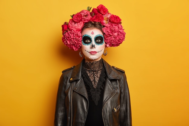 Free photo portrait of beautiful serious woman has creative vivid makeup, wears flower wreath, black clothes, tries be scarying, comes on halloween holiday party or day of dead