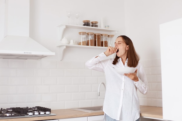 Free Photo portrait of beautiful sleepy dark haired young adult woman having breakfast in the kitchen, standing with raised arm, yawning, covering mouth with hand.