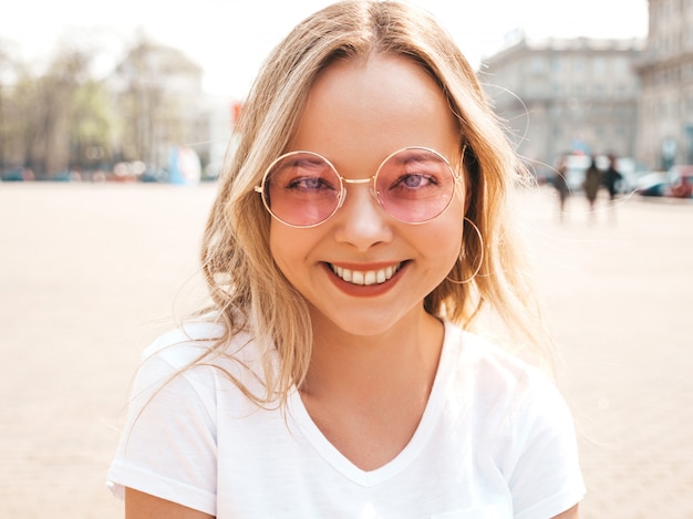 Portrait of beautiful smiling blond model dressed in summer hipster  clothes. Trendy girl posing in the street   in round sunglasses. Funny and positive woman having fun