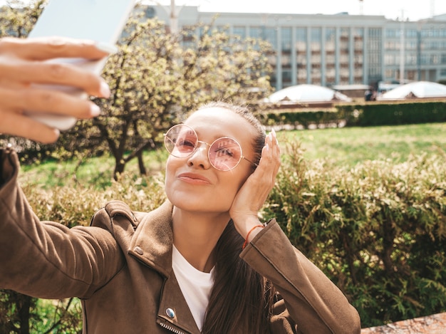 Free Photo portrait of beautiful smiling brunette girl in summer hipster jacket and jeans model taking selfie on smartphone