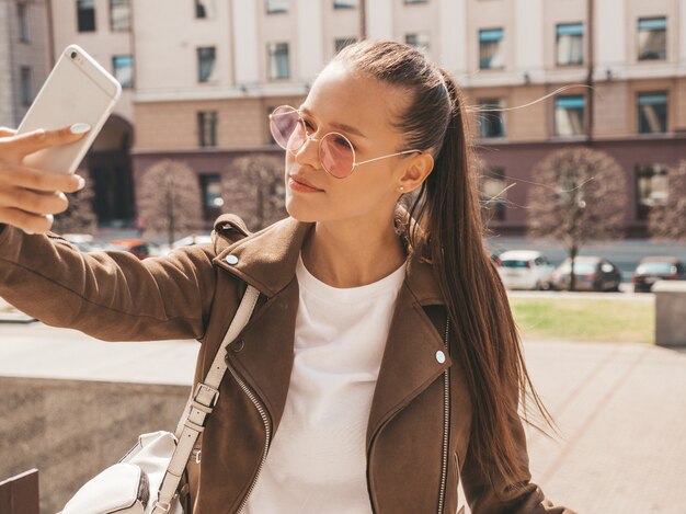 Portrait of beautiful smiling brunette girl in summer hipster jacket. Model taking selfie on smartphone.