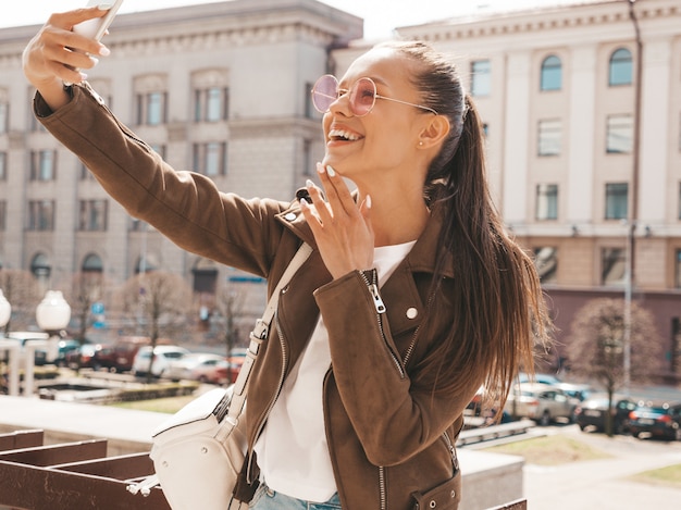 Portrait of beautiful smiling brunette girl in summer hipster jacket. Model taking selfie on smartphone.