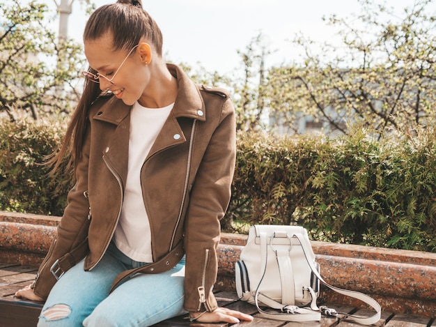 Free Photo portrait of beautiful smiling brunette model dressed in summer hipster jacket and jeans clothes  trendy girl sitting on the bench in the street   