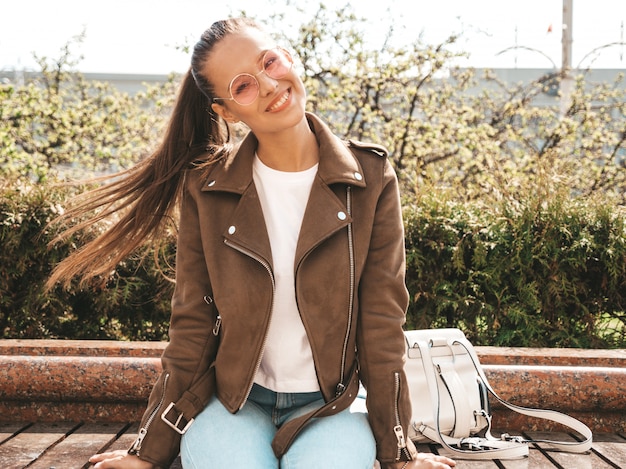 Free photo portrait of beautiful smiling brunette model dressed in summer hipster jacket and jeans clothes  trendy girl sitting on the bench in the street