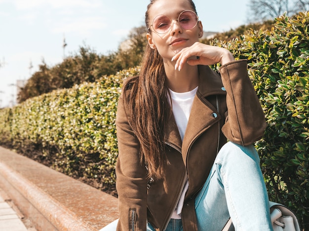 Free photo portrait of beautiful smiling brunette model dressed in summer hipster jacket and jeans clothes  trendy girl sitting on the bench in the street