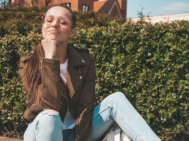 Free Photo portrait of beautiful smiling brunette model dressed in summer hipster jacket and jeans clothes  trendy girl sitting on the bench in the street   