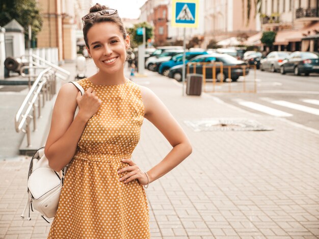 Portrait of beautiful smiling model dressed in summer yellow dress. Trendy girl posing in the street. Funny and positive woman having fun