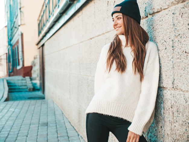 Portrait of beautiful smiling model. Female dressed in warm hipster white sweater and beanie. Posing in the street