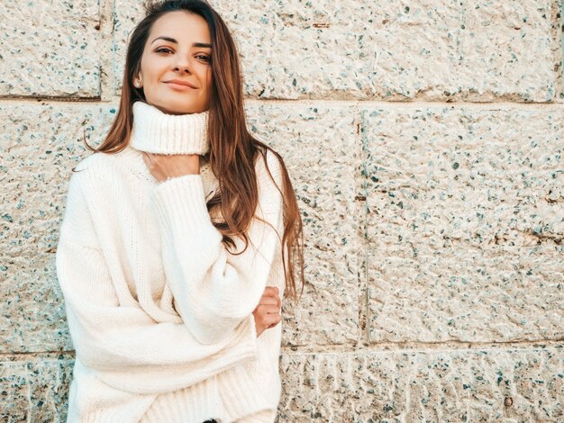 Portrait of beautiful smiling model. Female dressed in warm hipster white sweater. Posing near wall in the street