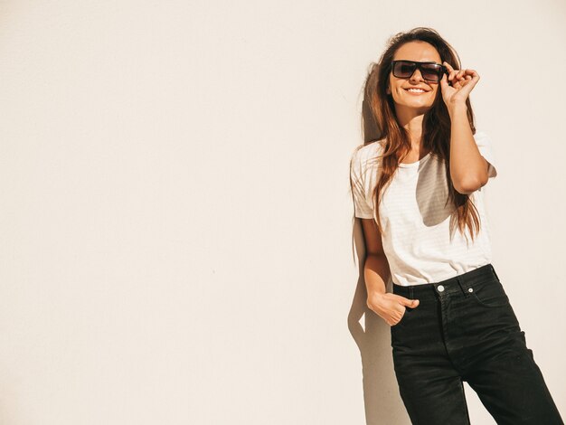 Portrait of beautiful smiling model in sunglasses. Female dressed in summer hipster white T-shirt and jeans. Trendy girl posing near wall in the street