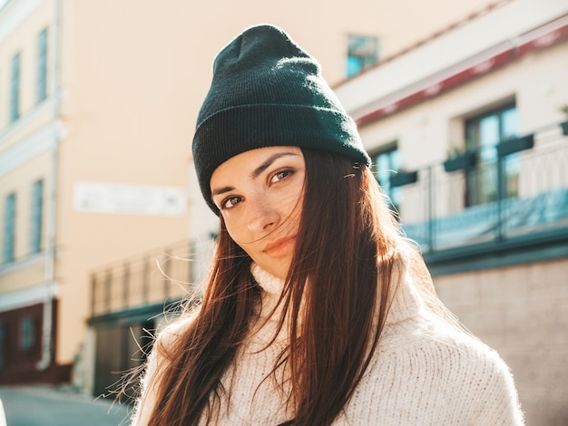 Portrait of beautiful smiling model. Woman dressed in warm hipster white sweater and beanie. She posing in the street