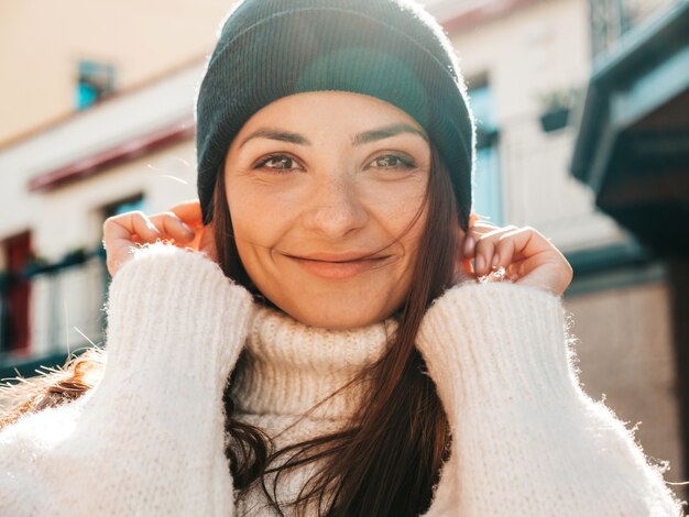 Portrait of beautiful smiling model. Woman dressed in warm hipster white sweater and beanie. She posing in the street