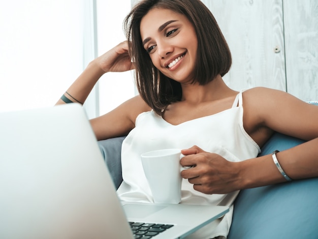 Portrait of beautiful smiling woman dressed in white pajamas. Carefree model sitting on soft bag chair and using laptop. 