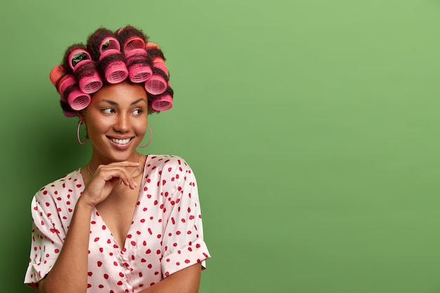 Portrait of beautiful smiling woman keeps hand under chin, poses with hair rollers in head for perfect curls, dressed in nightwear, prepares for special meeting, isolated on green wall, empty space