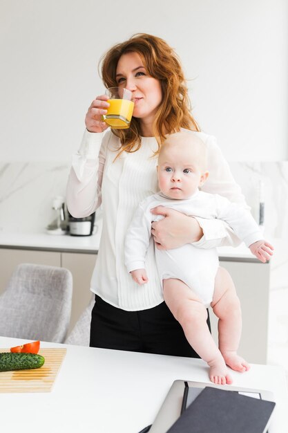 Portrait of beautiful smiling woman standing and holding her cute little baby while drinking juice and cooking on kitchen