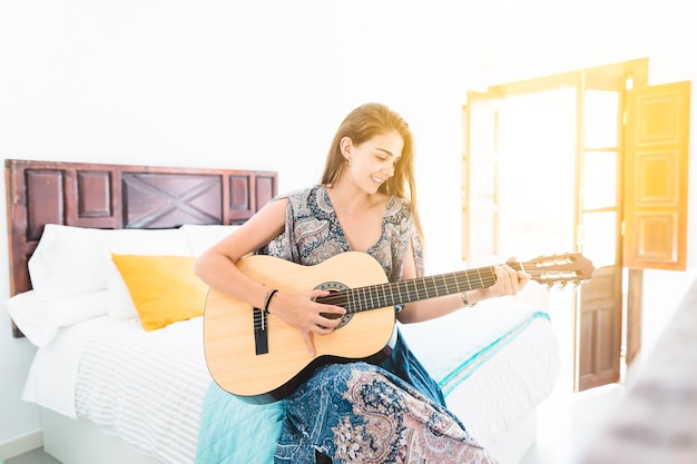 Free Photo portrait of beautiful teenage girl sitting on bed playing guitar