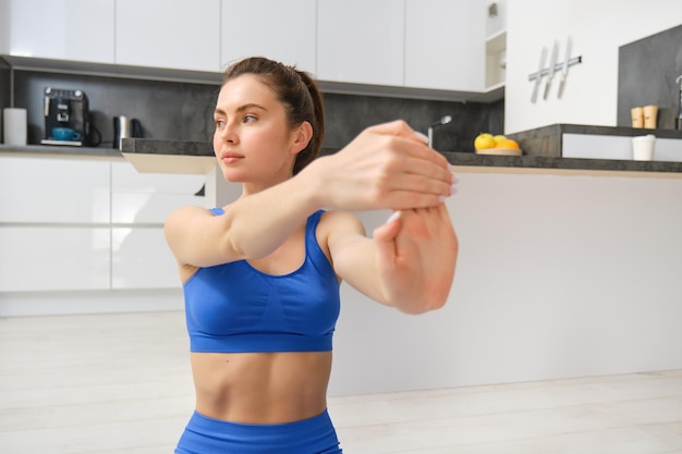 Portrait of beautiful woman does fitness training sits on floor near kitchen stretching her hands workout from home Copy space