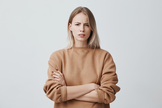 Free photo portrait of beautiful woman with blonde straight hair frowning her face in displeasure, wearing loose long-sleeved sweater, keeping arms folded. attractive young woman in closed posture.