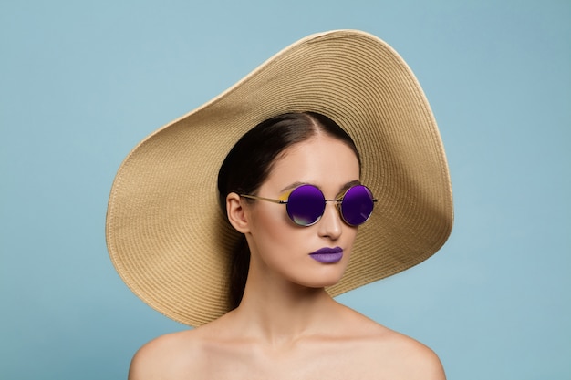 Free photo portrait of beautiful woman with bright make-up, hat and sunglasses on blue studio background. stylish and fashionable make and hairstyle. colors of summer. beauty, fashion and ad concept. serious.