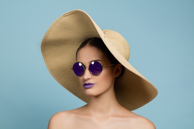 Free photo portrait of beautiful woman with bright make-up, hat and sunglasses on blue studio