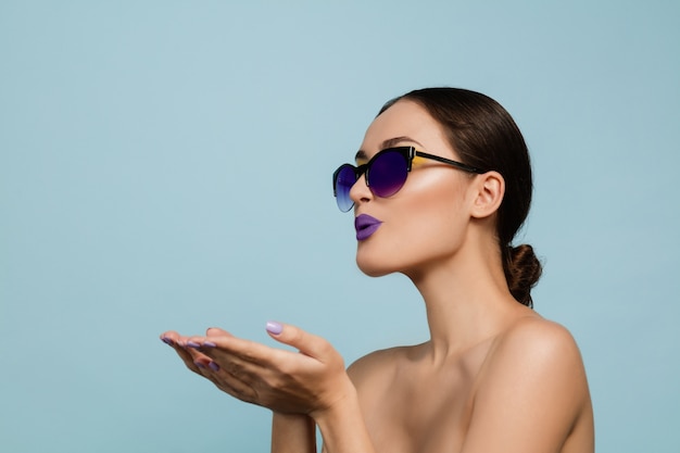 Portrait of beautiful woman with bright make-up and sunglasses on blue studio background. Stylish, fashionable make and hairstyle. Colors of summer. Beauty, fashion and ad concept. Sending kisses.