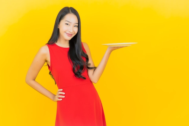 Free photo portrait beautiful young asian woman smile with empty plate dish on yellow wall