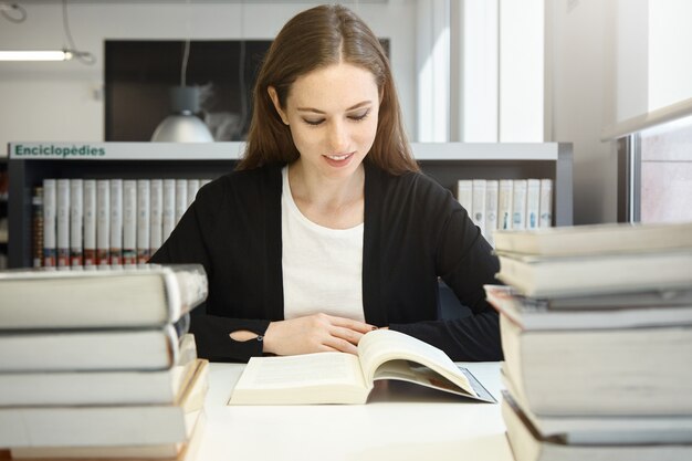 Portrait of beautiful young brunette female professor wearing black jacket reading manual or textbook, smiling, preparing for lecture in university, sitting at library in front of stacks of books