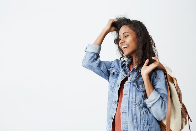 Portrait of beautiful young dark-skinned female with wavy dark hair wearing casual red T-shirt and denim jacket with backpack on her shoulder smiling pleasantly. Trendy hipster woman having good mood