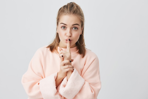 Portrait of beautiful young fair-haired female wearing stylish pink sweatshirt asking to keep silence or not to tell her secrets, having serious look, holding finger on her lips. Body language