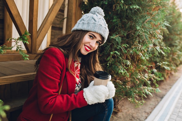 Free photo portrait beautiful young girl with long hair in red coat sitting on wooden stairs outdoor. she has grey knitted hat, white gloves, holds coffee and smiling .