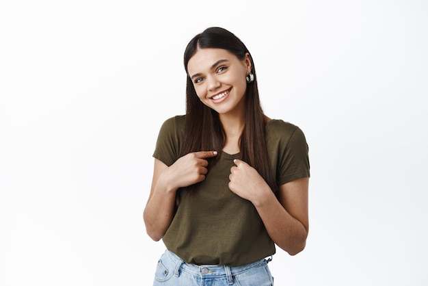 Portrait of beautiful young healthy woman in tshirt pointing at herself and smiling shy selfpromoting asking to pick her standing against white background