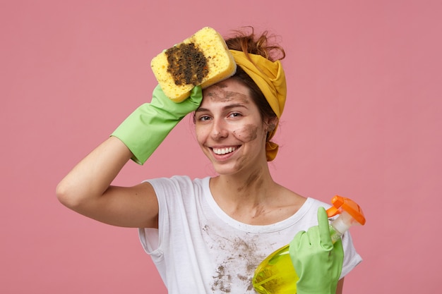 Portrait of beautiful young housewife with dirty clothes and face holding mop and washing spray holding hand on head looking tired but happy to finish work. Tired cute woman doing house cleaning