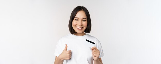 Portrait of beautiful young modern asian woman showing credit card and thumbs up recommending contactless payment standing over white background