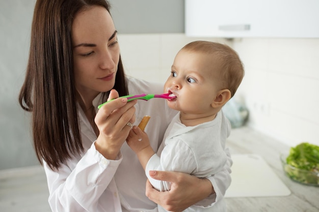 Portrait of beautiful young mother feeding her newborn child with porridge Son eating from spoon and looking aside with funny expression