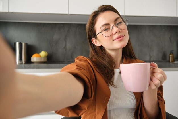 Free Photo portrait of beautiful young woman in glasses takes selfie with cup of tea sitting in kitchen holding