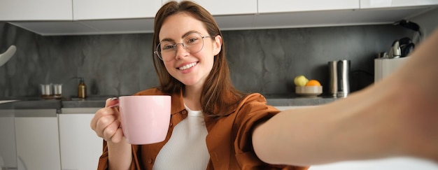 Free Photo portrait of beautiful young woman in glasses taking selfie in kitchen with cup of morning coffee