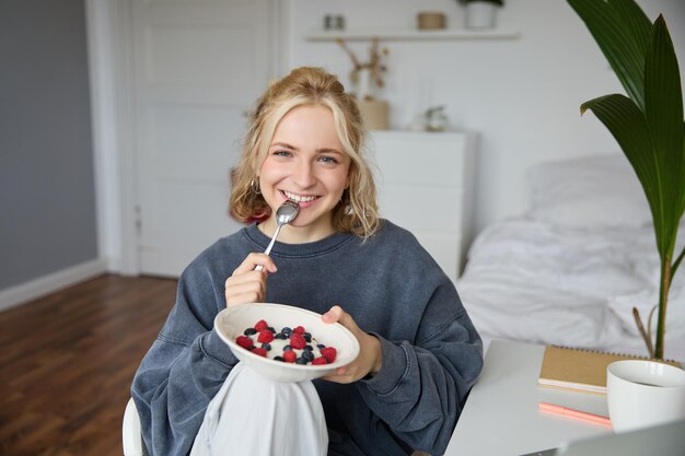 Portrait of beautiful young woman in a room eating breakfast holding bowl with dessert and a spoon