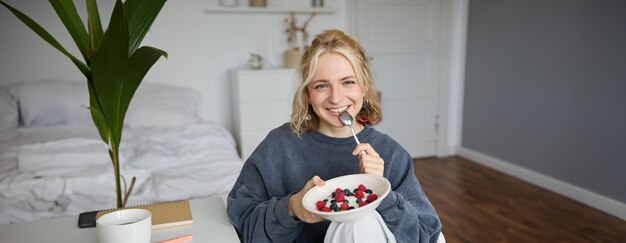 Portrait of beautiful young woman in a room eating breakfast holding bowl with dessert and a spoon