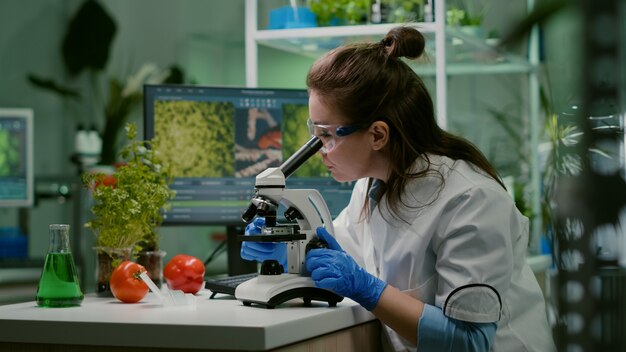 Portrait of biologist scientist in white coat working in expertise laboratory looking into microscope analyzing organic gmo leaf