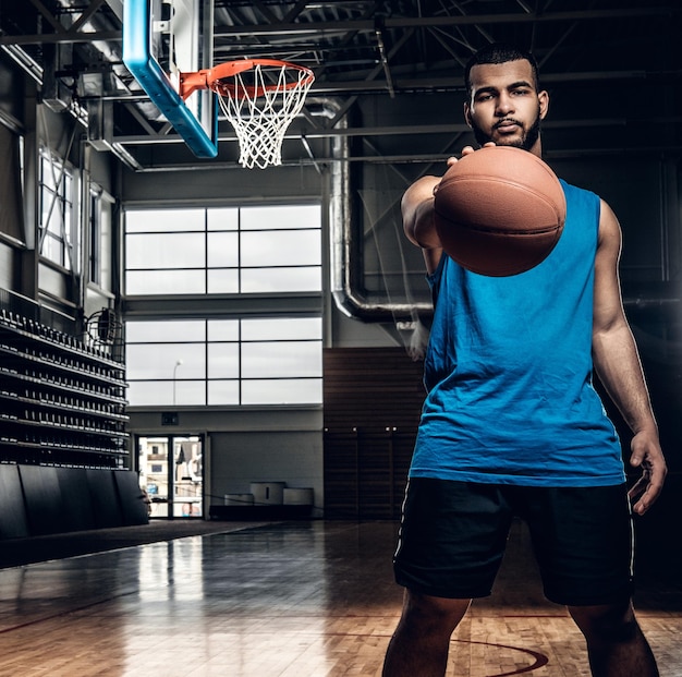 Portrait of Black basketball player holds a ball over a hoop in a basketball hall.