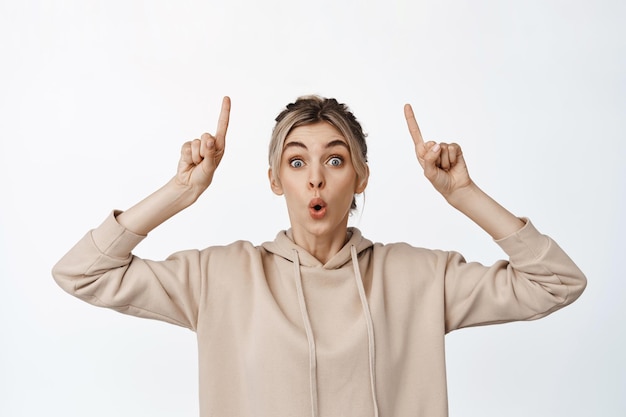 Free Photo portrait of blond girl with surprised face pointing fingers up and say wow impressed by sale or discount banner stands against white background