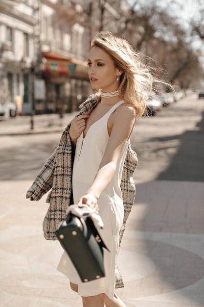 Portrait of blonde beautiful curly woman in stylish silk white dress and tweed oversized jacket holding black leathered handbag and walking outdoors