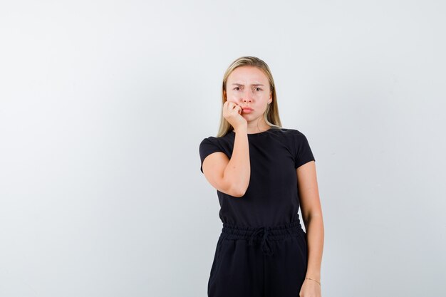 Portrait of blonde lady keeping hand on cheek in black dress and looking gloomy front view