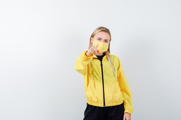 Portrait of blonde lady pointing at camera in tracksuit, mask and looking serious front view