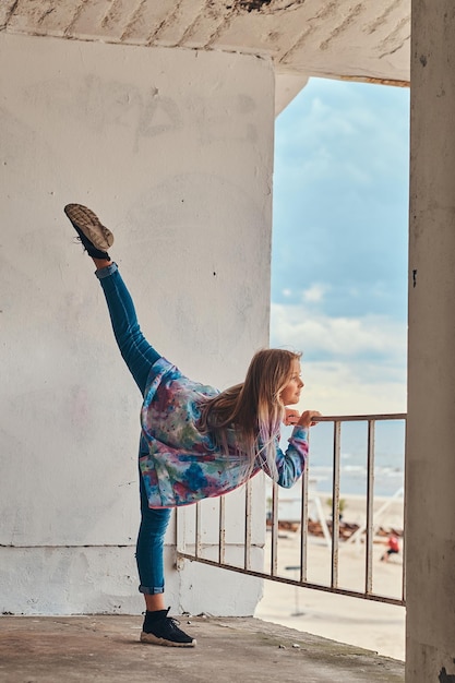 Free photo portrait of a blonde schoolgirl dressed in trendy clothes posing with raised leg while leaning on a guardrail against a sea coast.