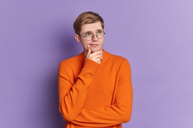 Free photo portrait of blue eyed guy ponders on something stands in thoughtful pose holds chin concentrated into distance wears casual orange jumper