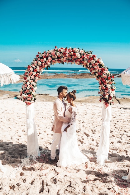 Free photo portrait of bride and groom posing near wedding tropical arch on the beach behind blue sky and sea. wedding couple