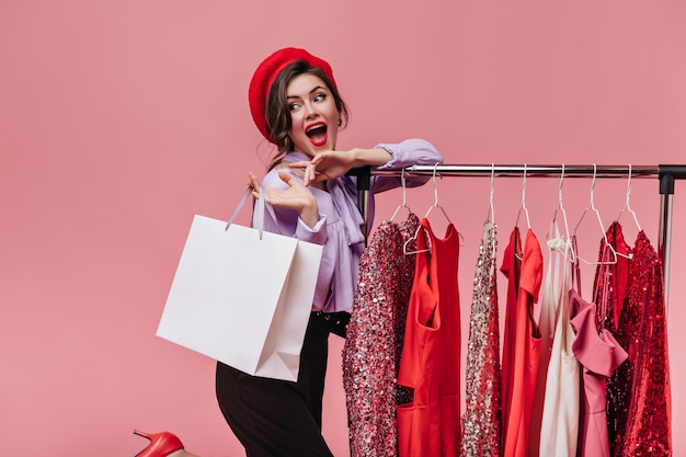 Portrait of bright lady with red lipstick posing happily with package while shopping on pink background.