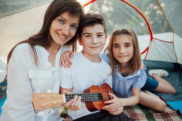 Portrait of brother and sister holding ukulele sitting with their mother in tent