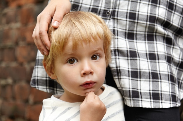 Free Photo portrait of brown-eyed boy and his mother. five-years old kid with blond hair standing quietly. his loving mom standing near, stroking his head. curious facial expression.